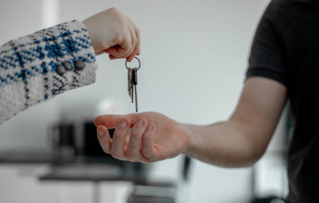Close-up photo of real estate saleswoman in plaid jacket handing keys to new condominium to tall male client in black T-shirt after signing contract